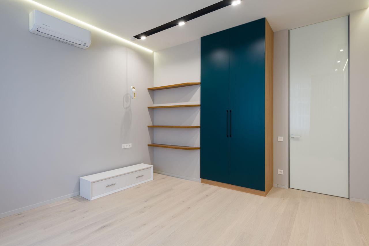 Wooden wardrobe and shelves near white wall in modern room with parquet floor and bright lamps
